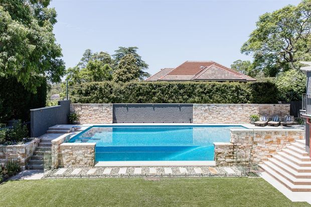 A landscaped backyard swimming pool with a raised stone-clad pool edge and feature wall, surrounded by pale stone paving, clipped hedges, mature trees and a manicured lawn. A tiled pitched roof of the house is visible beyond the garden, with steps and a deck area at right.