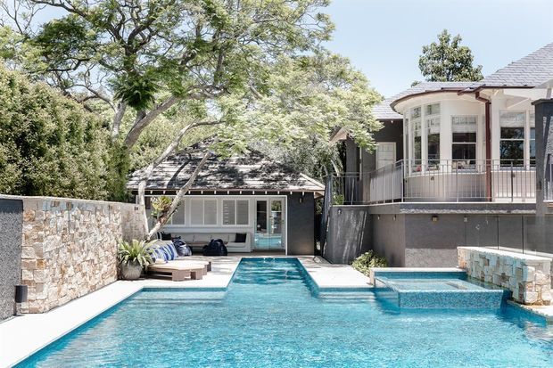 Exterior view of a substantial residential property with a large swimming pool and raised spa in the foreground. The pool courtyard is framed by stone retaining walls, mature trees, a low dark-clad pool or guest pavilion, and a main house with a curved glazed bay, balcony railing, and tiled pitched roof.