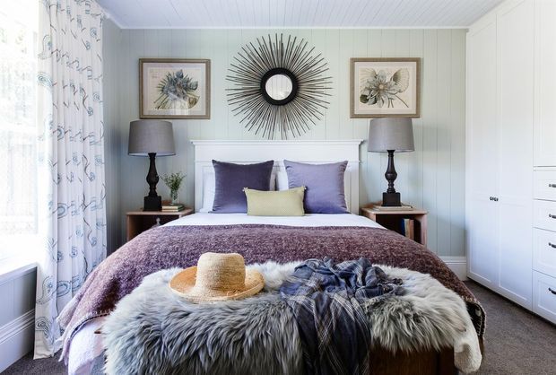 A bright, inviting bedroom with white vertical wall panelling and a matching white paneled ceiling. The bed is layered with lavender bedding, a textured mauve throw, a pale yellow accent cushion, and a large grey faux-fur throw at the foot. Dark timber bedside tables and lamps flank the bed, while a large sunburst mirror and two framed botanical artworks decorate the feature wall. White built-in wardrobes occupy the right side, and patterned curtains frame the window on the left.