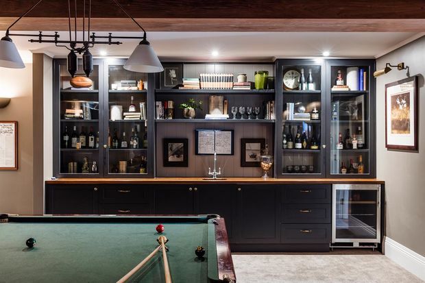 A refined home bar and billiards room with extensive dark built-in cabinetry, glass-front display shelves, a central sink, under-counter wine refrigerator, and a green-felt pool table in the foreground. Warm timber ceiling beams, pendant lighting, framed artwork, and brass-toned hardware create a classic, intimate atmosphere.
