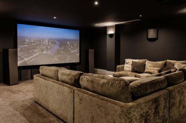 A dark, dedicated home cinema with a large projection screen, wall-mounted speakers, recessed ceiling lighting, and a substantial U-shaped sectional sofa arranged for viewing. The room has deep brown-black walls and ceiling, plush taupe upholstery, and carpeted flooring, creating a moody and immersive atmosphere.
