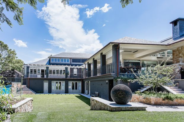 A spacious landscaped courtyard and lawn framed by a large multi-level residence. The home combines light rendered walls, dark timber or metal detailing, broad covered terraces, extensive glazing, stone masonry, and low-pitched tiled or metal roofing. A large spherical sculpture sits on a stone plinth in the foreground, with outdoor stairs, planted beds, and a bright blue sky completing the scene.