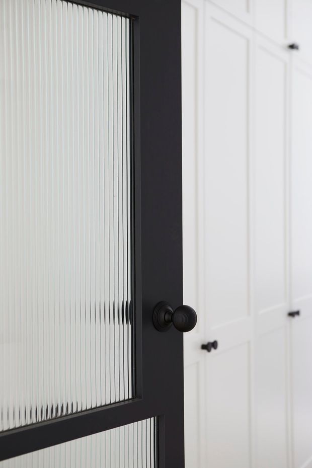 Close-up interior detail of a black-framed reeded glass door with a dark round knob, positioned in front of white paneled cabinetry or doors with black hardware. The image emphasises contrasting monochrome joinery and textured glazing.