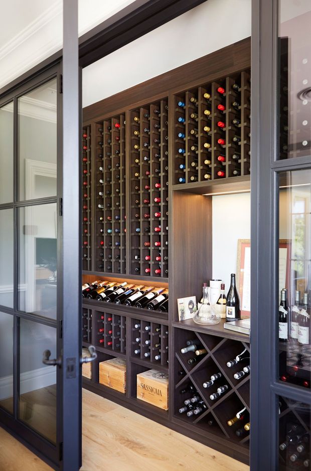 A compact, custom-built wine storage room viewed through dark-framed glazed doors. Floor-to-ceiling dark timber wine racks hold numerous bottles, with lower diamond-grid storage, display shelving, a timber drawer unit, and a small tasting or serving shelf. Pale timber flooring, white walls, and black-framed glazing create a refined, tailored interior.