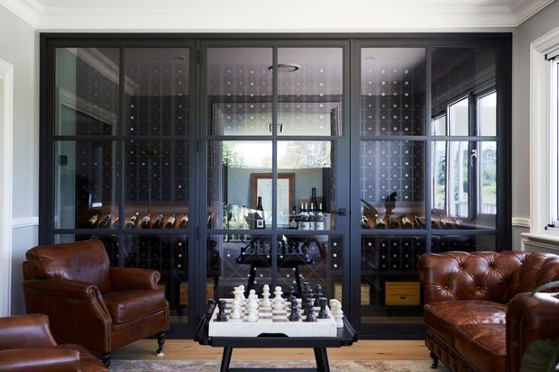 A refined home lounge featuring a large built-in glass-fronted wine display and bar cabinet in dark joinery, flanked by richly upholstered brown leather armchairs and a tufted leather sofa. A chess table sits in the foreground, with classic white wall panelling, ornate ceiling cornices, and views through reflective glazing adding depth.