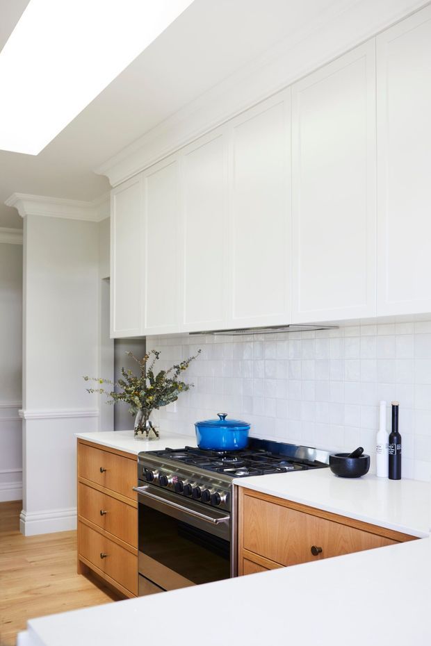 Bright, refined kitchen with white raised-panel upper cabinetry, warm timber drawer and base cabinetry, white countertops, and a glossy white tiled splashback. A large stainless-steel range cooker with a vivid blue Dutch oven forms the focal point. Decorative wall mouldings, cornices, and a large skylight add a traditional architectural character, while timber flooring brings warmth.