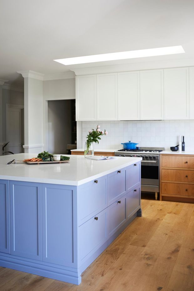 Bright, refined kitchen with a large pale blue panelled island, white shaker-style upper cabinetry, white tiled splashback, natural timber flooring, and warm timber lower drawers. A freestanding stainless-steel range cooker sits along the rear wall beneath the cabinetry, with a skylight or large ceiling light opening above.
