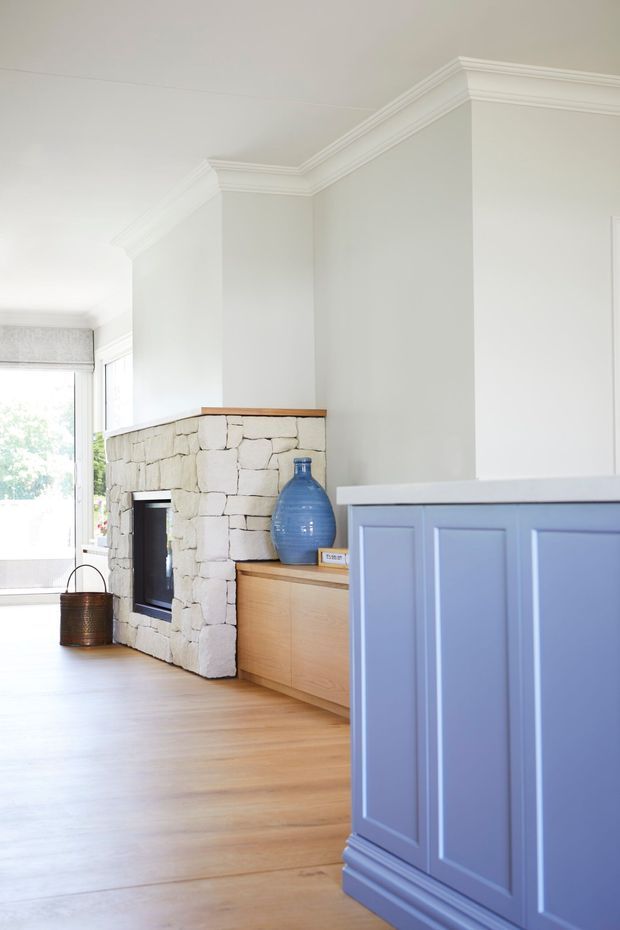 Bright, refined living area with pale walls, ornate white crown moulding, light timber flooring, a white irregular-stone fireplace surround, and warm timber built-in cabinetry. A blue-painted paneled cabinet or kitchen island occupies the foreground, with a large blue ceramic vessel adding colour. Natural light enters through tall glazing at the rear.