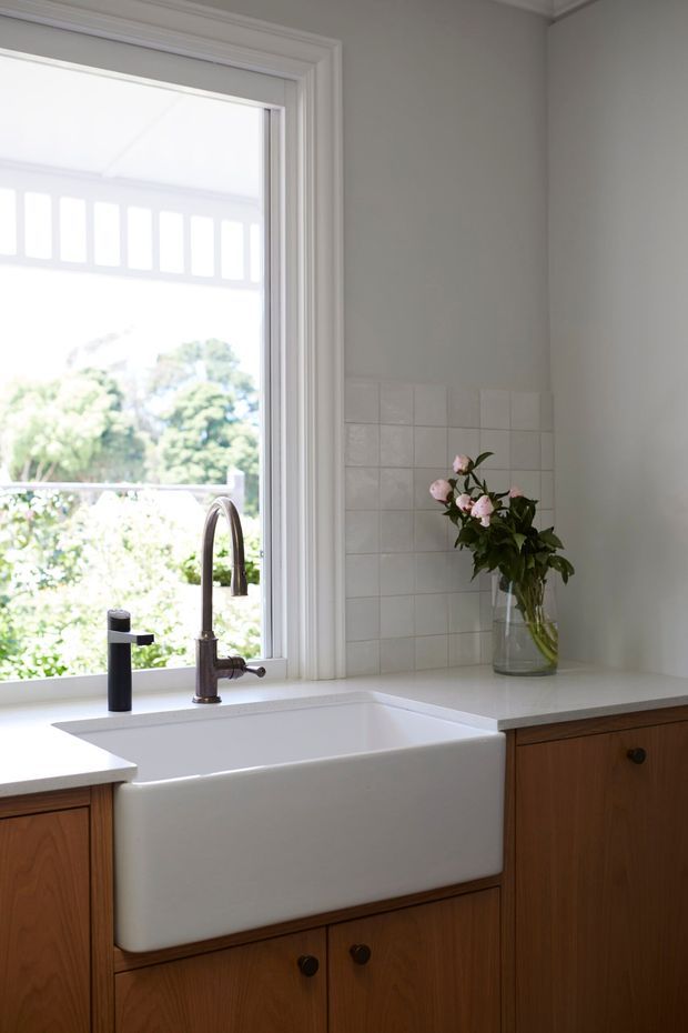 Bright kitchen sink area with a deep white farmhouse apron-front sink set into white countertops, dark metal tapware, and warm timber cabinetry. A white square-tile splashback and traditional layered window trim frame the view to greenery outside. A glass vase with flowers sits on the benchtop.