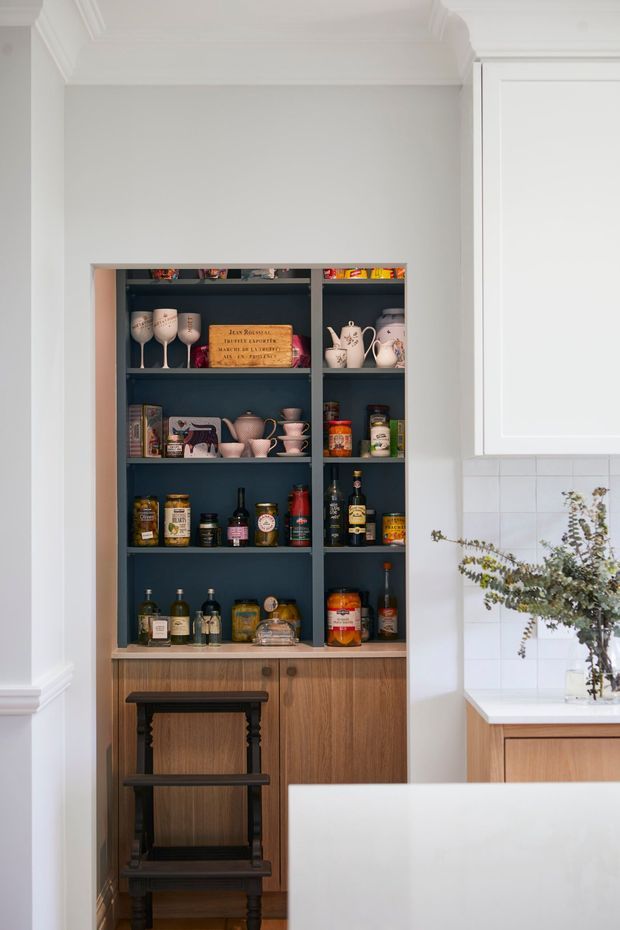 A refined kitchen interior featuring a dark blue open display pantry or bar cabinet with walnut-toned lower cabinetry, stocked with glassware, ceramics, bottles, and pantry items. White shaker-style cabinetry, pale tiled backsplash, a light stone benchtop, decorative ceiling cornices, and a dark timber step stool create a classic, tailored look.