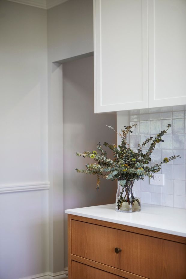 A close-up view of a refined kitchen or kitchenette corner featuring white raised-panel overhead cabinetry, warm timber drawer cabinetry, a white benchtop, and a pale square-tile splashback. Soft grey walls with decorative wall moulding and a vase of eucalyptus-style foliage create a calm, classic interior.