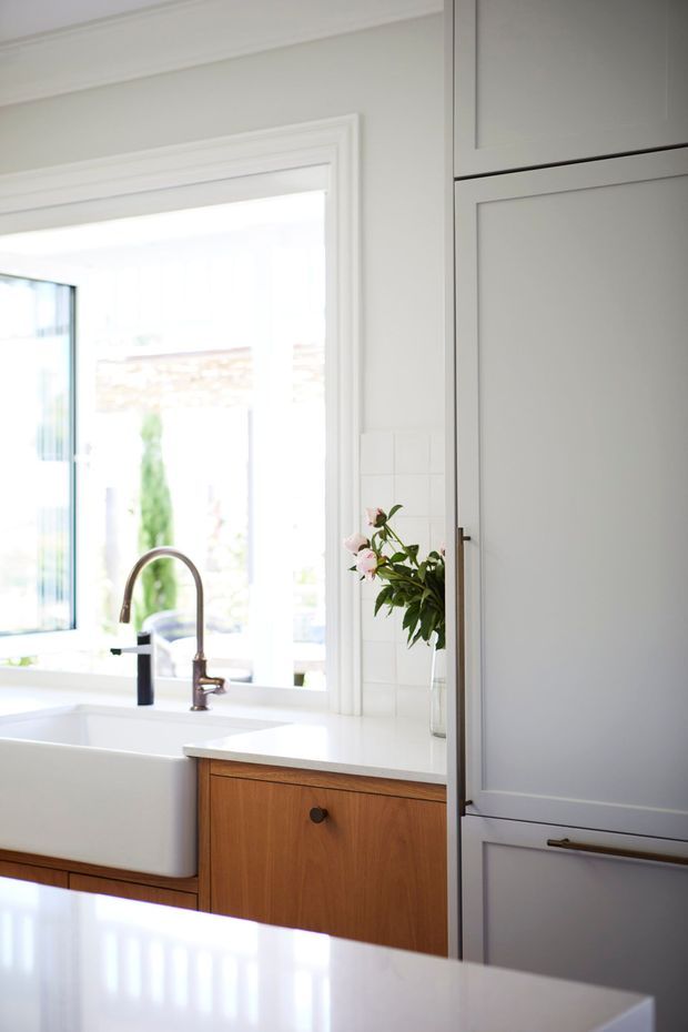 Bright, refined kitchen interior featuring a white farmhouse sink beneath a large window, a curved brushed-metal faucet, warm timber cabinetry, white benchtops, white tiled splashback, and a tall pale-grey built-in cabinet. Traditional moulded trim surrounds the opening, while a vase of pink flowers adds a small decorative accent.