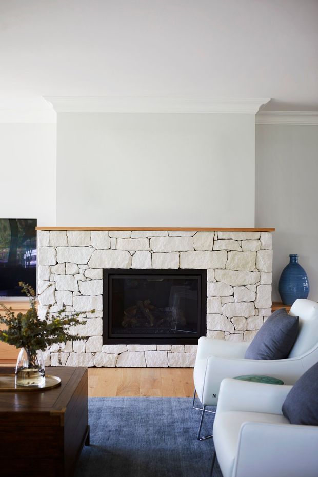 Bright, refined living room centered on a large white rough-stone fireplace with a black inset firebox and timber mantel. White armchairs with dark blue cushions face the fireplace, while a dark timber coffee table, blue rug, ceramic vase, television, and light timber flooring complete the space. Traditional wall and ceiling mouldings contrast with the contemporary furnishings.