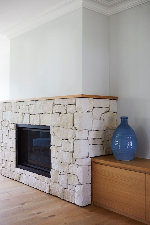 Bright, refined living room detail featuring a substantial white/cream irregular stone fireplace with a black inset firebox. A warm timber mantel and low built-in timber cabinetry extend to the right, complemented by light timber flooring, white walls, and decorative crown moulding. A large blue glass vessel provides a contrasting accent.