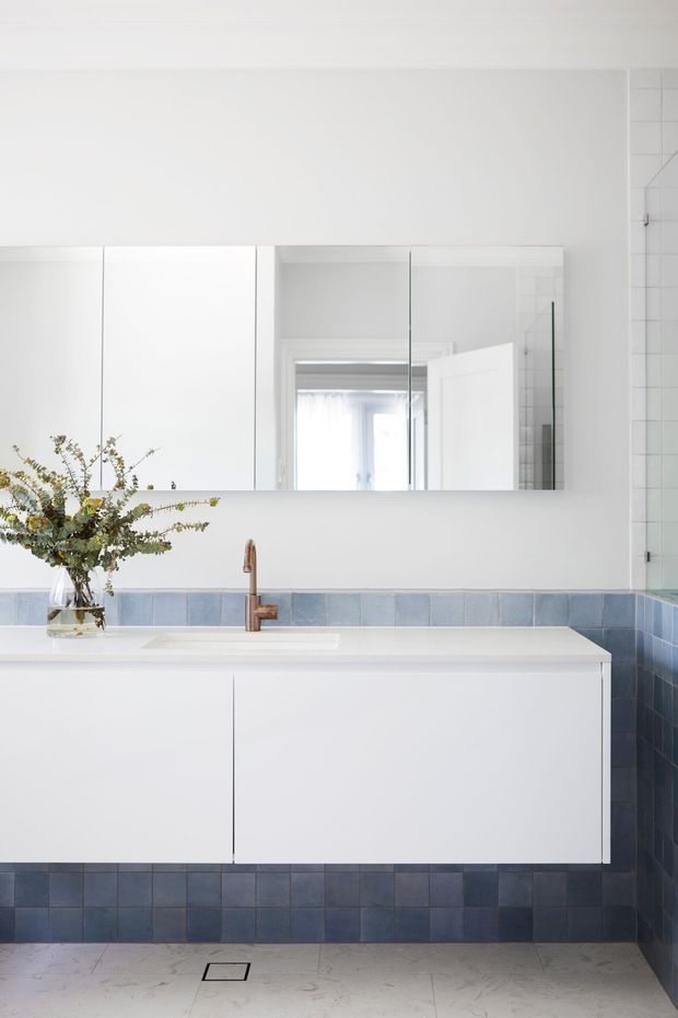 Bright contemporary bathroom vanity with a wall-mounted white cabinet, integrated basin, mirrored storage above, and a bronze-toned tap. Blue square wall tiles form a backsplash and lower wall treatment, continuing into the shower area at right, while pale stone-look floor tiles and white walls create a clean, light interior.