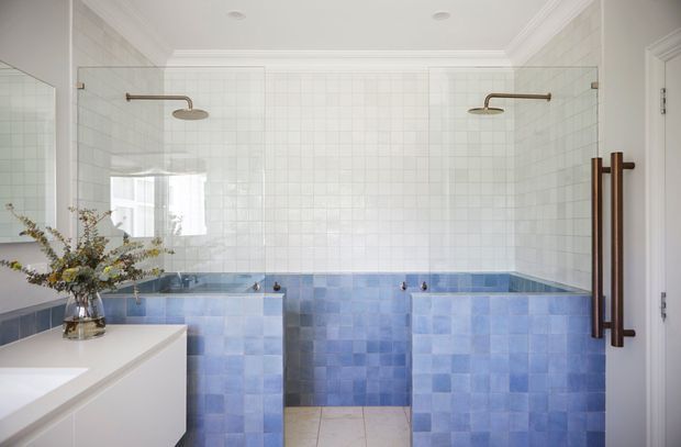Bright, refined bathroom with twin open shower bays, white grid-pattern wall tiles, and blue tiled half-height partitions and wall surfaces. The room includes a white vanity, large mirror, recessed ceiling lights, and warm bronze shower and door hardware.