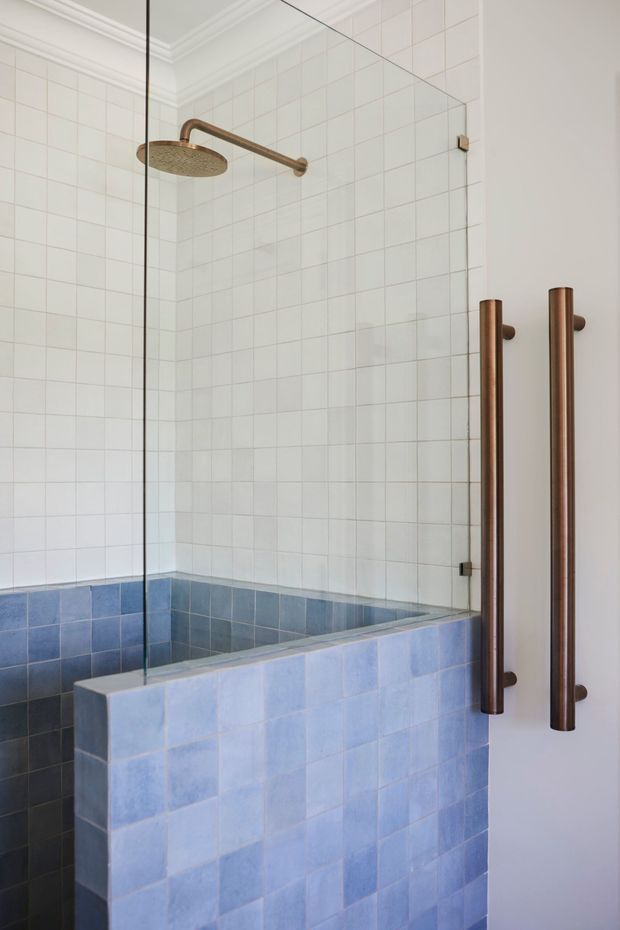 A refined bathroom shower enclosure with a frameless glass screen, white square wall tiles, and a lower wall clad in blue ceramic tiles. The shower features a large round bronze showerhead and matching bronze vertical door handles. White decorative ceiling cornices add a traditional architectural detail.