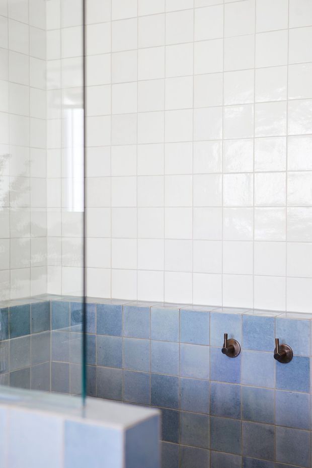 A bright tiled bathroom shower or bath area with a frameless glass screen. The walls are clad in glossy white square tiles above a band of blue ceramic tiles, with dark bronze wall-mounted plumbing fixtures. The space has a clean, simple, slightly retro character.
