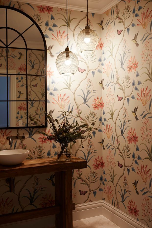 A richly decorated powder room with full-height botanical wallpaper featuring flowers, leaves, birds and butterflies. A dark timber console supports a white vessel basin, with a large black-framed arched mirror and two woven pendant lights. Ornate ceiling and skirting details complete the warm, characterful interior.
