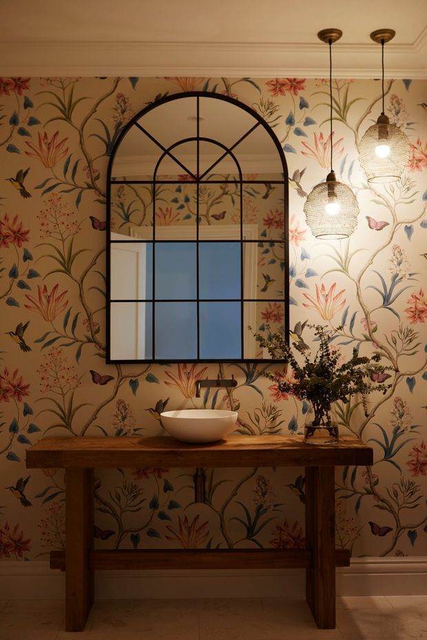 A richly decorated bathroom vanity area with a rustic solid timber console, white vessel basin, floral botanical wallpaper, and a large black arched industrial-style mirror. Two woven pendant lights hang from a detailed moulded ceiling, creating a warm, atmospheric interior.