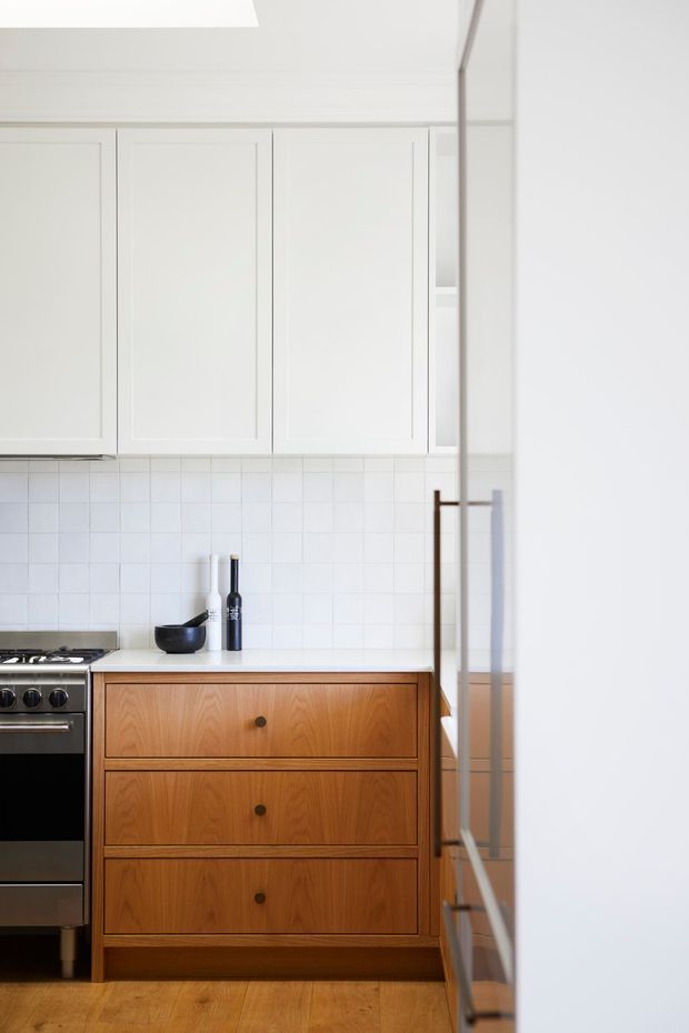 A bright, compact kitchen with white recessed-panel wall cabinetry, a white tiled splashback, white benchtop, and warm timber drawer cabinetry. A stainless-steel freestanding oven sits to the left, with a small black bowl and dark bottle on the counter. Timber flooring and a large reflective glazed surface at right complete the minimalist composition.