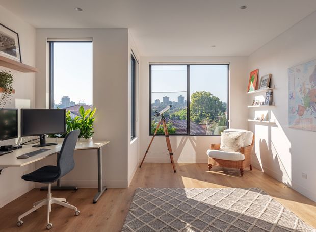 Bright contemporary home office with a built-in or wall-adjacent desk, dual computer monitors, ergonomic office chair, indoor plants, large windows, a telescope, framed artwork, open display shelving, and a comfortable armchair. Warm timber flooring, white walls, and abundant natural light create an airy workspace overlooking greenery and a distant urban skyline.