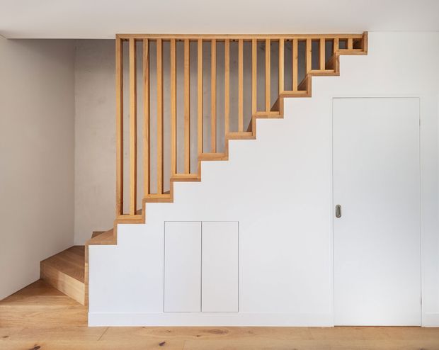 A minimalist residential stairway viewed side-on, with warm timber stair treads and vertical timber balusters forming a diagonal open railing. The stair structure and under-stair wall are painted white, with integrated flush storage doors beneath the stairs and a tall flush white door to the right. Light timber flooring and neutral plaster walls create a clean, understated interior.