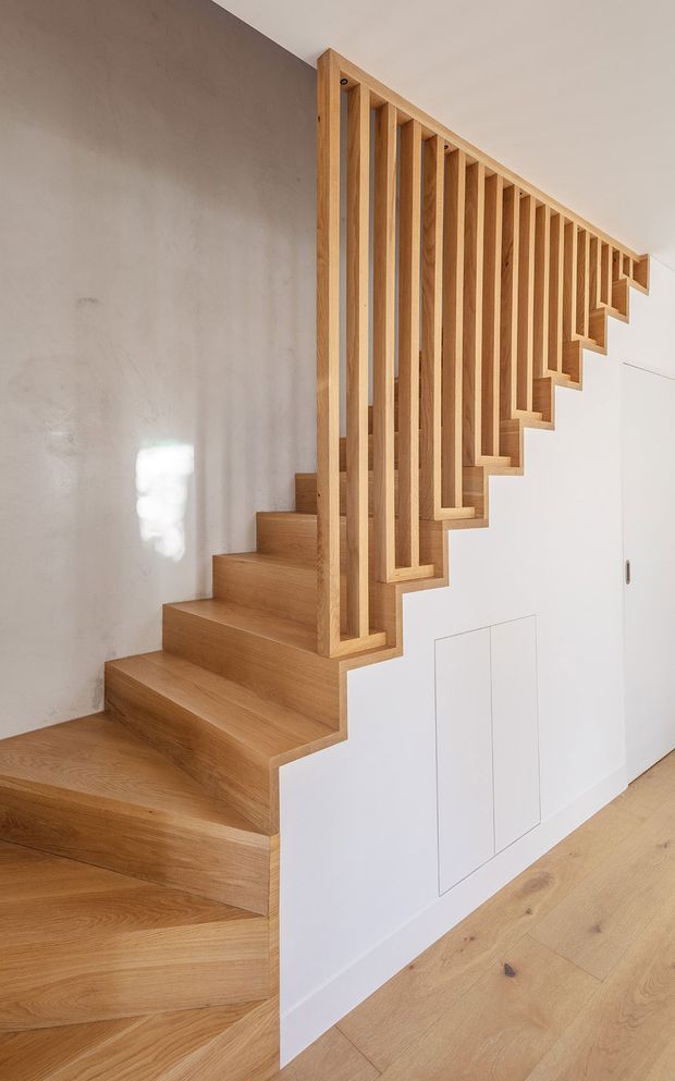 Interior view of a contemporary timber staircase with broad oak-toned treads, matching vertical slatted balustrade, and integrated white under-stair cabinetry. The staircase is set against a smooth warm-grey plaster wall, with pale timber flooring extending into the adjoining space. Clean lines, concealed storage doors, and natural wood detailing create a warm minimalist composition.