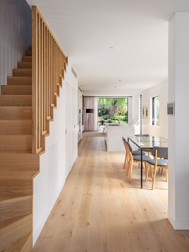 A bright, contemporary open-plan residential interior viewed from an entry or hallway. A timber staircase with a vertical timber slat balustrade runs along the left side, while pale timber flooring leads toward a dining area and white kitchen/living space at the rear. Large windows and glazing provide views to a leafy garden, and the room is furnished with a glass dining table and light upholstered seating. The palette is predominantly white and light natural timber, creating an airy, refined atmosphere.