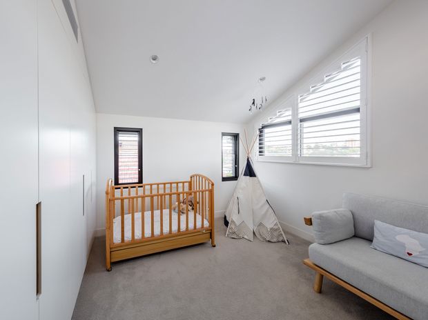 Bright, compact nursery with a natural timber cot, grey upholstered sofa, children's teepee, built-in white wardrobe cabinetry, soft grey carpet, and white plantation-shutter windows. The room has a clean, calming and contemporary family-oriented interior.