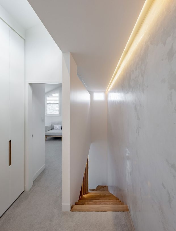A narrow, minimalist stairway viewed from an upper hallway, with warm timber treads descending beside a smooth white textured wall. A concealed linear LED strip illuminates the stair wall, while white built-in doors and a carpeted landing lead toward a softly furnished room with a window.