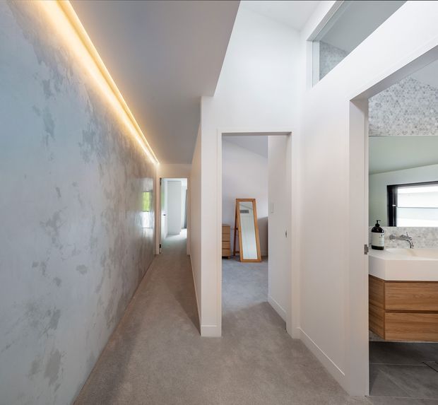 A bright contemporary residential hallway with a long textured feature wall and concealed linear LED lighting. The carpeted corridor opens toward a bedroom or dressing area, while a partially visible bathroom with a timber vanity, white basin, dark tiled floor, and patterned wall tile is seen through an open doorway. White walls, crisp door frames, and high-level glazing create an airy, refined interior.
