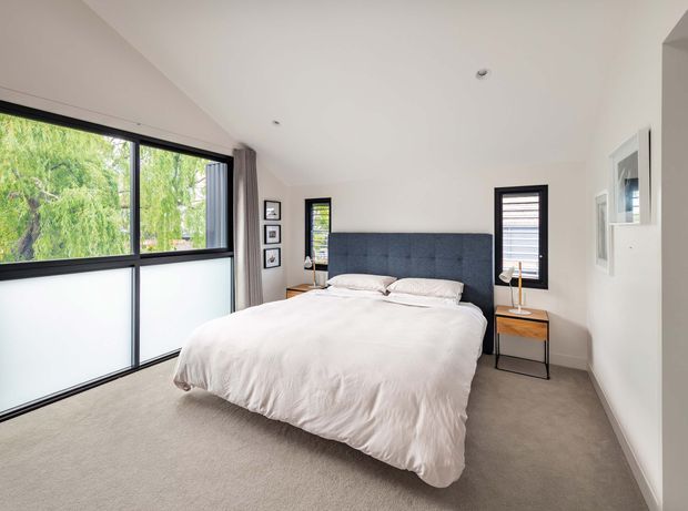 Bright contemporary bedroom with a large upholstered bed and deep blue-grey headboard, white bedding, beige carpet, slim timber bedside tables, and black-framed windows. Frosted lower glazing provides privacy while greenery is visible through the upper panes. The room has a clean, understated fit-out with a sloping ceiling and framed wall art.