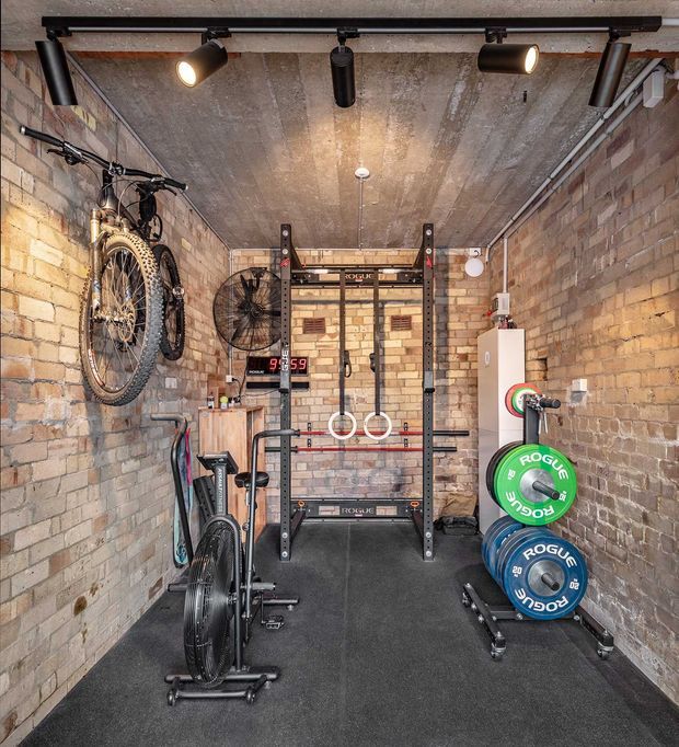Compact brick-walled home gym with an exposed concrete ceiling, black rubber flooring, track lighting, wall-mounted bicycles, an Olympic lifting rack with gymnastic rings, bumper plates, an air bike, and other workout equipment. The utilitarian industrial structure is softened by warm lighting and exposed brick.