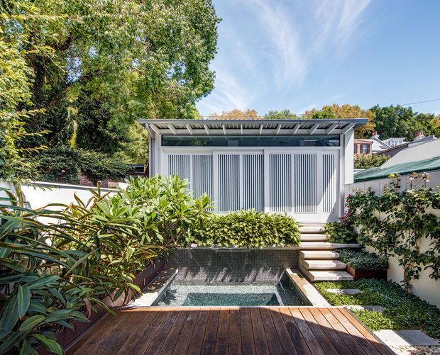 A landscaped residential courtyard viewed from a timber deck, featuring a small rectangular plunge pool, planted garden beds, pale rendered boundary walls, and a compact contemporary pavilion or pool house with a shallow metal roof and vertical slatted screening. The setting is leafy and established, with steps connecting the deck, pool edge, and pavilion.