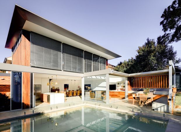A modern two-storey residence viewed from beside a swimming pool. The home features broad floor-to-ceiling glazing, timber wall cladding, extensive horizontal external louvres, a projecting flat roof, and a sheltered outdoor entertaining area with timber screening and built-in seating. The bright open-plan interior is visible through the glazing, while the pool occupies the foreground.