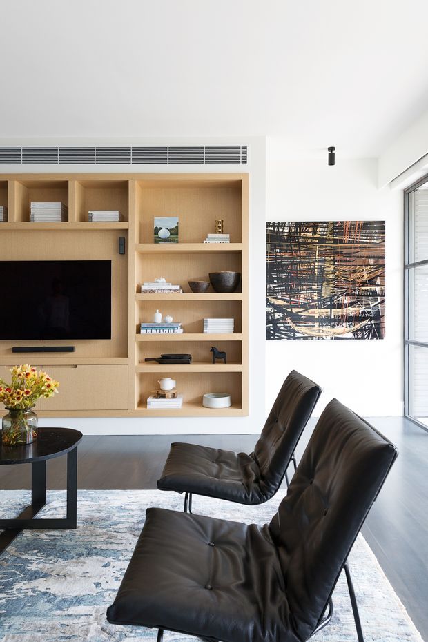 Bright contemporary living room with a built-in light timber entertainment wall and open shelving, black television, sculptural objects, and a large abstract artwork. Two black leather lounge chairs sit over a patterned area rug beside a dark coffee table. Full-height glazing and white walls create an airy, refined interior.