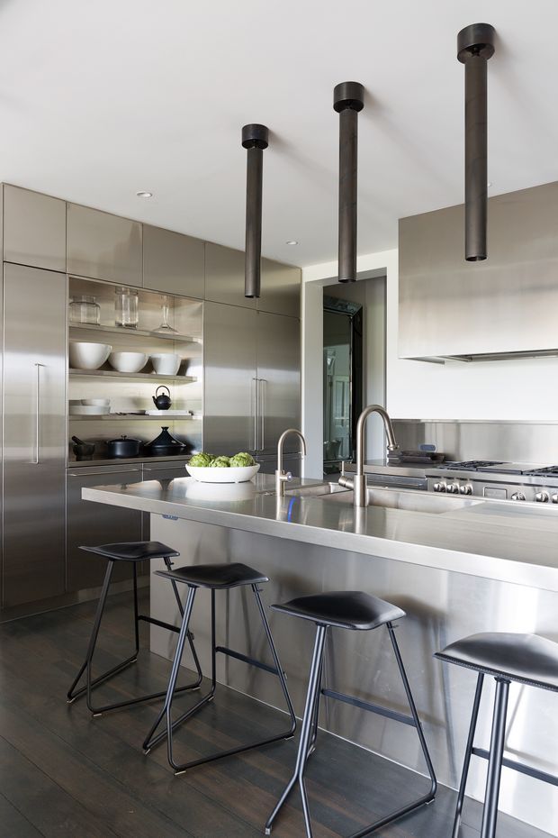 A sleek contemporary kitchen with a large stainless-steel island and integrated sink, gas cooktop, and extensive grey cabinetry. Open shelving displays crockery and glassware, while four black stools line the island. Three dark cylindrical pendant lights hang from a smooth white ceiling, and dark timber flooring adds contrast.