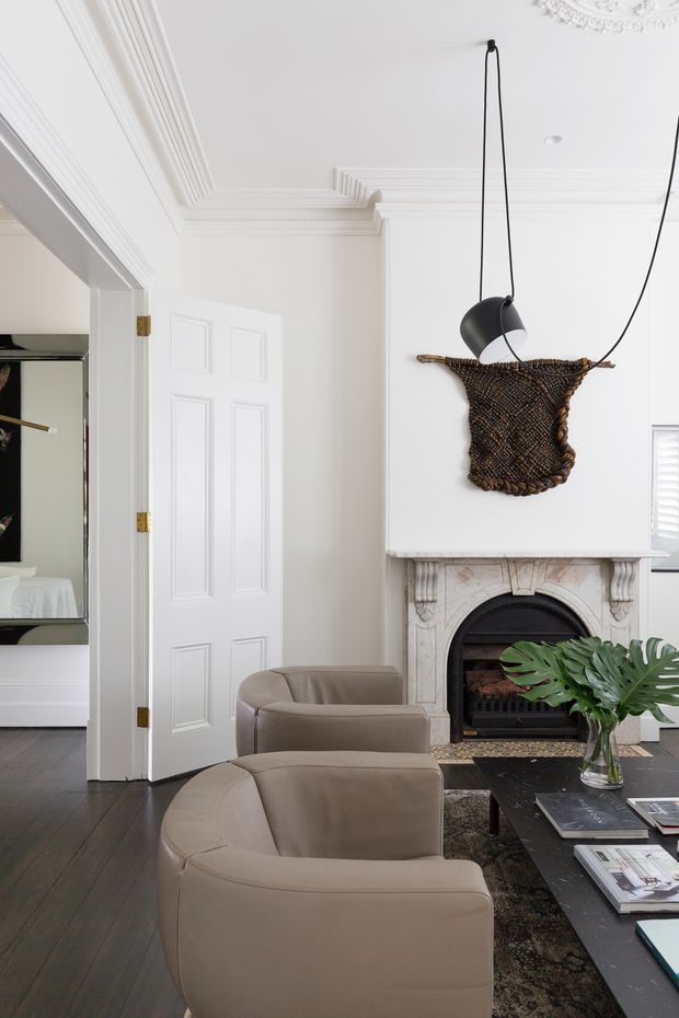 Elegant, bright living room with white walls, elaborate decorative ceiling cornices, dark timber flooring, and a carved marble fireplace with a black firebox. Two rounded taupe upholstered armchairs face a low dark coffee table with books and a glass vase of large green leaves. A sculptural black pendant light hangs above an organic woven wall artwork, while a white paneled door and glimpsed adjacent room sit to the left.