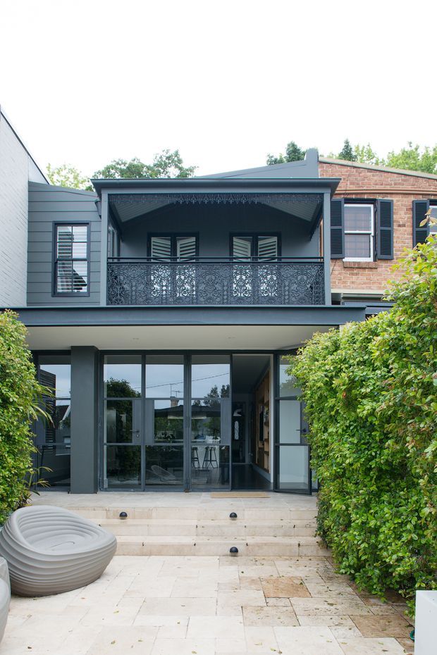 Rear exterior view of a two-storey urban home with a contemporary dark-grey ground-floor extension, large glazed doors, pale stone-paved terrace and steps, dense hedge planting, and an upper-level covered balcony with ornate perforated metal balustrade. The original upper facade combines grey cladding with exposed red brick and dark shutters.