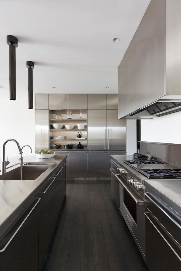 A sleek contemporary galley kitchen with dark cabinetry, stainless-steel appliances, integrated floor-to-ceiling storage, open shelving, and a large stainless-steel sink and benchtop. The space has a white ceiling, dark timber flooring, minimalist cylindrical pendant lights, and a restrained monochromatic palette.
