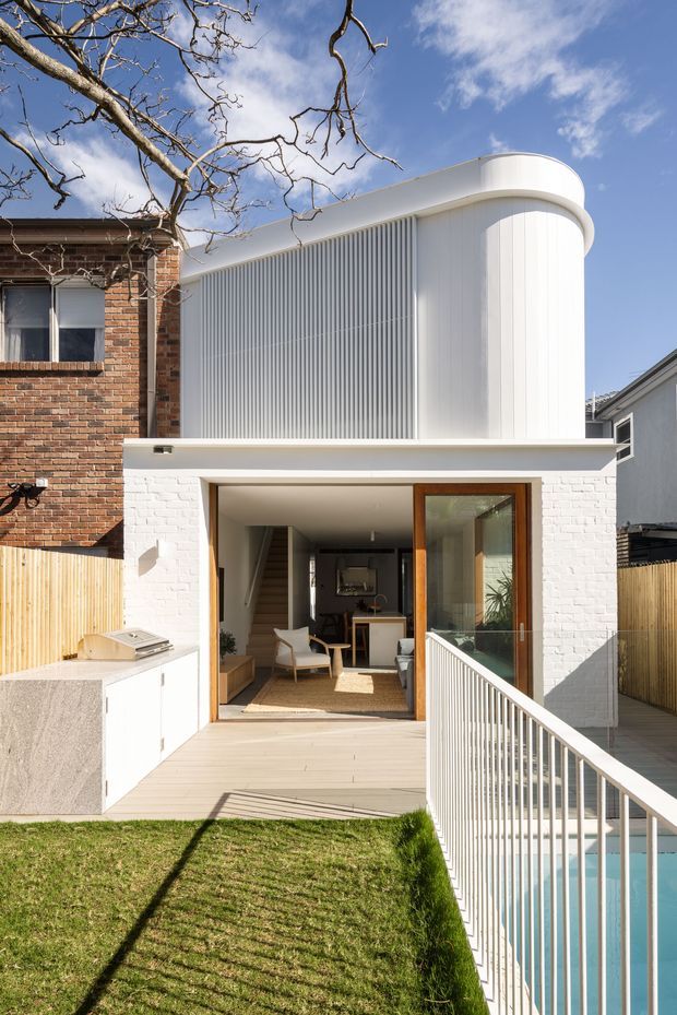 A bright rear exterior view of a contemporary residential addition. A white-painted brick ground floor opens onto a timber deck and narrow lawn, with a pool and white balustrade at right. Above, a distinctive curved white upper volume features vertical ribbed screening and rounded roofline detailing. A contrasting older red-brick neighbouring or retained structure is visible on the left, while large timber-framed glazing connects the interior living area to the deck.