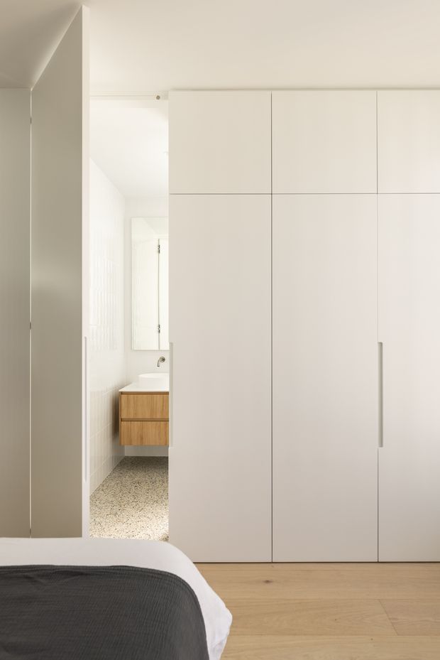 A minimalist bedroom with a partially visible bed in the foreground, pale timber flooring, and a large wall of smooth white built-in wardrobes with concealed-style vertical handles. An open doorway reveals a compact bathroom with a timber vanity, white basin, mirror, and terrazzo-like flooring. The space is bright, restrained, and highly streamlined.