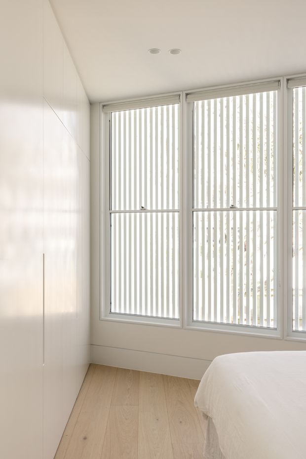 Bright, minimalist bedroom corner with a partially visible white bed, floor-to-ceiling built-in white cabinetry, pale timber flooring, and large windows fitted with vertical white blinds. The room has a clean, airy appearance and recessed ceiling downlights.