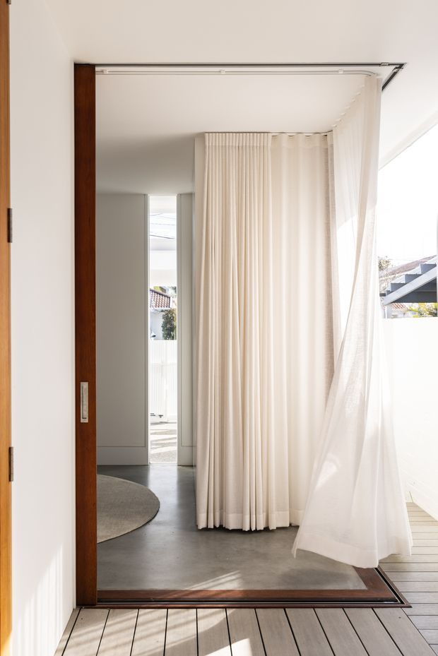 Bright minimalist interior transition space with a large timber-framed glazed opening, polished concrete floor, and full-height sheer cream curtains. The opening leads toward an entry or hallway, with a timber deck visible outside and strong natural light filtering through the curtains.