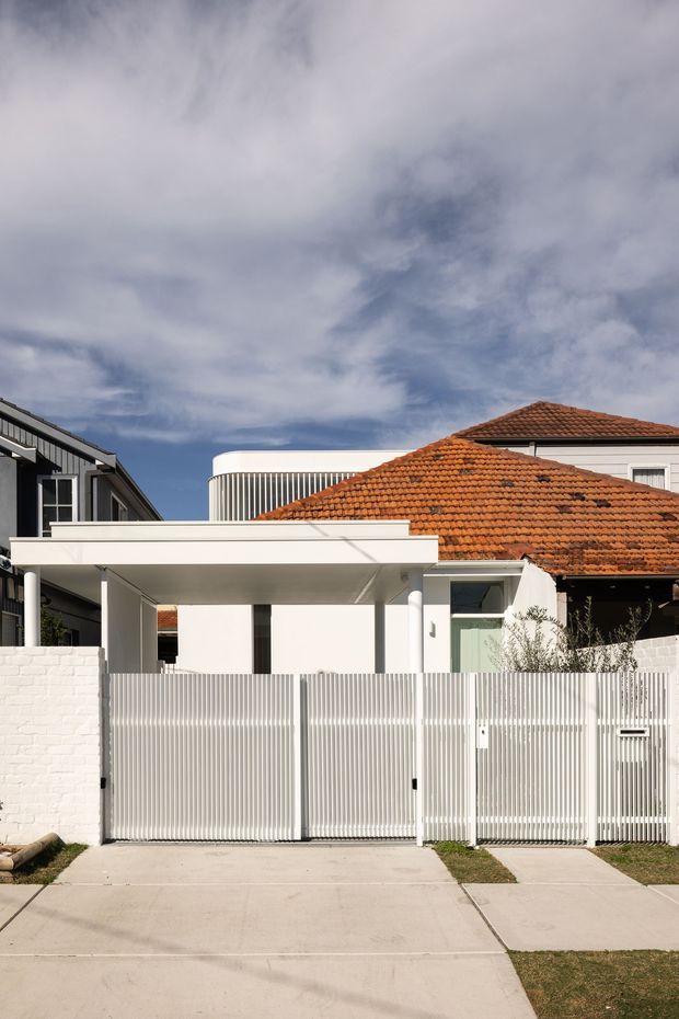 Street-facing view of a contemporary white residential facade with a flat-roofed carport, vertical slatted metal fencing and gate, white rendered walls, and a prominent terracotta tiled pitched roof from the adjoining or retained structure. The home sits in a suburban streetscape with a concrete driveway and sidewalk.