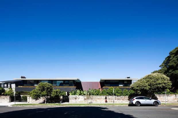 Street-facing view of a low-rise contemporary residential building composed of two flat-roofed dark-clad wings connected by a central reddish feature volume. Extensive horizontal glazing, balconies, stone boundary walls, mature landscaping, and a parked car are visible under a clear blue sky.