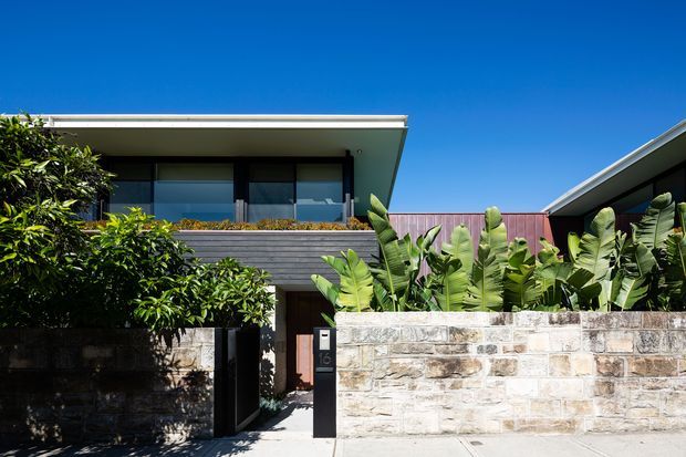 Bright exterior view of a contemporary two-storey residence with a flat-roofed upper level, expansive dark-framed glazing, horizontal dark cladding, and a sheltered entry gate. A pale stone masonry boundary wall and lush tropical planting define the front garden, while a second sloping roof volume is visible at right.