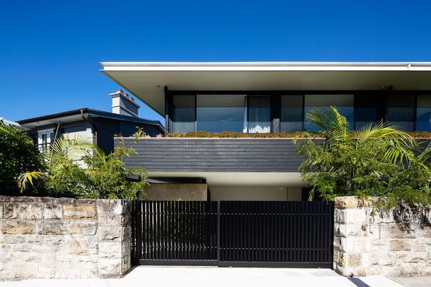Street-facing exterior view of a contemporary multi-storey residence with a broad flat roof, projecting upper-level balcony, dark horizontal cladding, extensive glazing, black metal privacy fencing, and pale natural-stone boundary walls. Tropical planting frames the entrance and the facade is strongly lit against a clear blue sky.