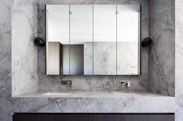 A contemporary bathroom vanity viewed straight on, featuring a wide double-sink stone benchtop, mirrored wall cabinet, dark wall-mounted sconces, and dramatic grey-veined marble or stone slabs covering the walls and counter. Dark fluted cabinetry is visible below the vanity.